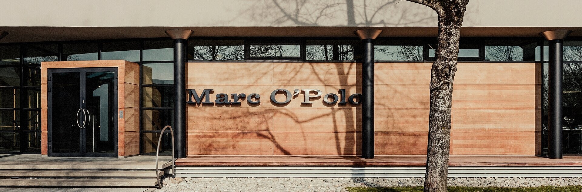 Gebäude Marc O Polo mit Schriftzug vorn, Holzfassade, Baum und Straße davor