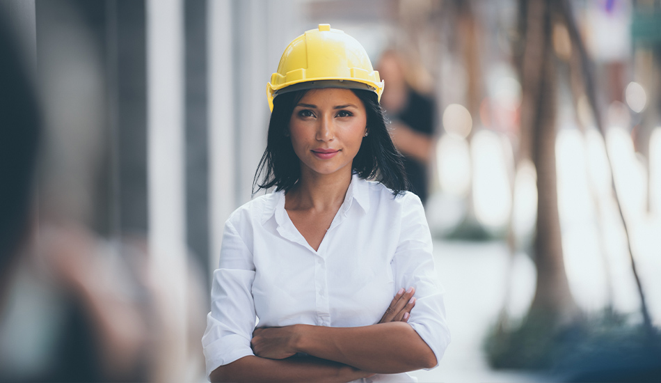 Woman standing in front of construction site with helmet on head and arms crossed