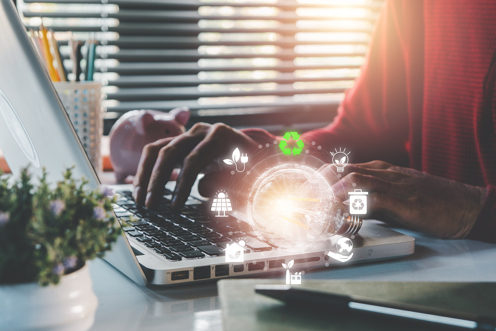 Frau sitzt am Schreibtisch und tippt auf Laptop, über dem Laptop schweben digitale Symbole wie Glühbirne, Solaranlage und Recycling-Pfeile zum Thema Nachhaltigkeit.