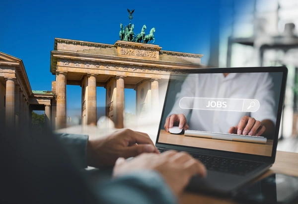 Im Vordergrund ein Mann, der an einem Laptop nach Jobs sucht, im Hintergrund Brandenburger Tor