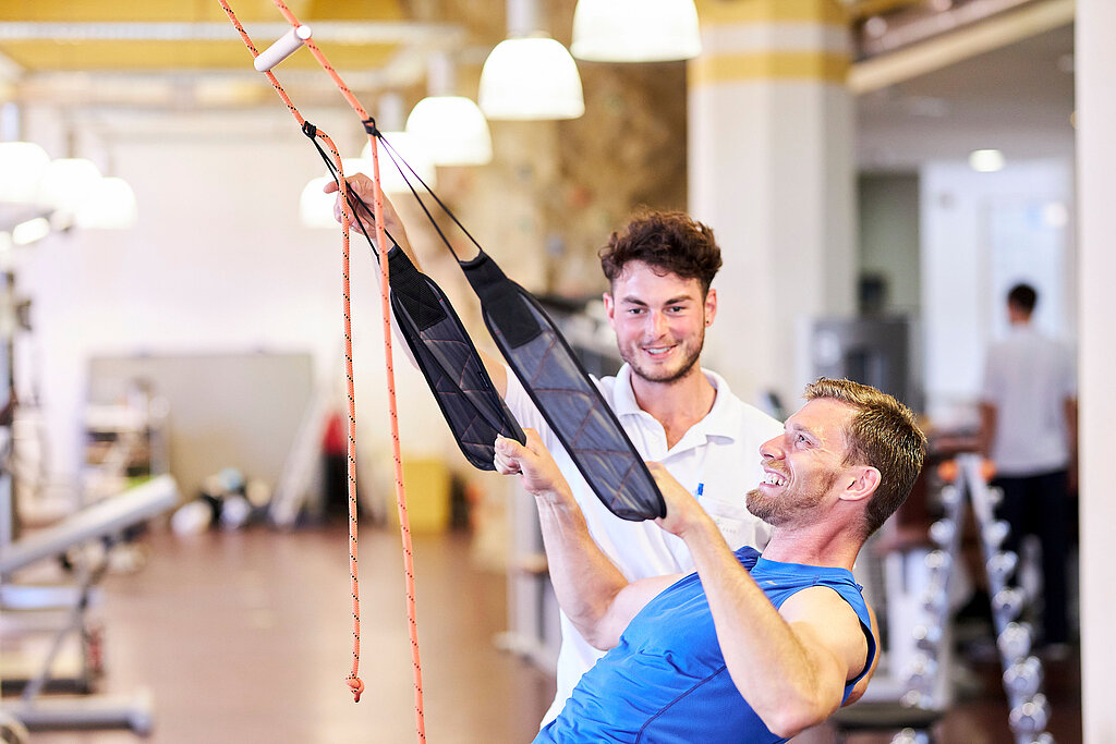 2 Männer in einem Fitnessstudio, einer trainiert seine Oberarme, der andere gibt Hilfestellung