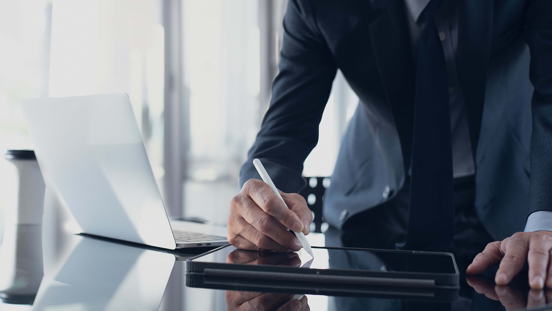 A man in a suit is drawing something on his tablet, which is lying on his desk. Next to it is a laptop