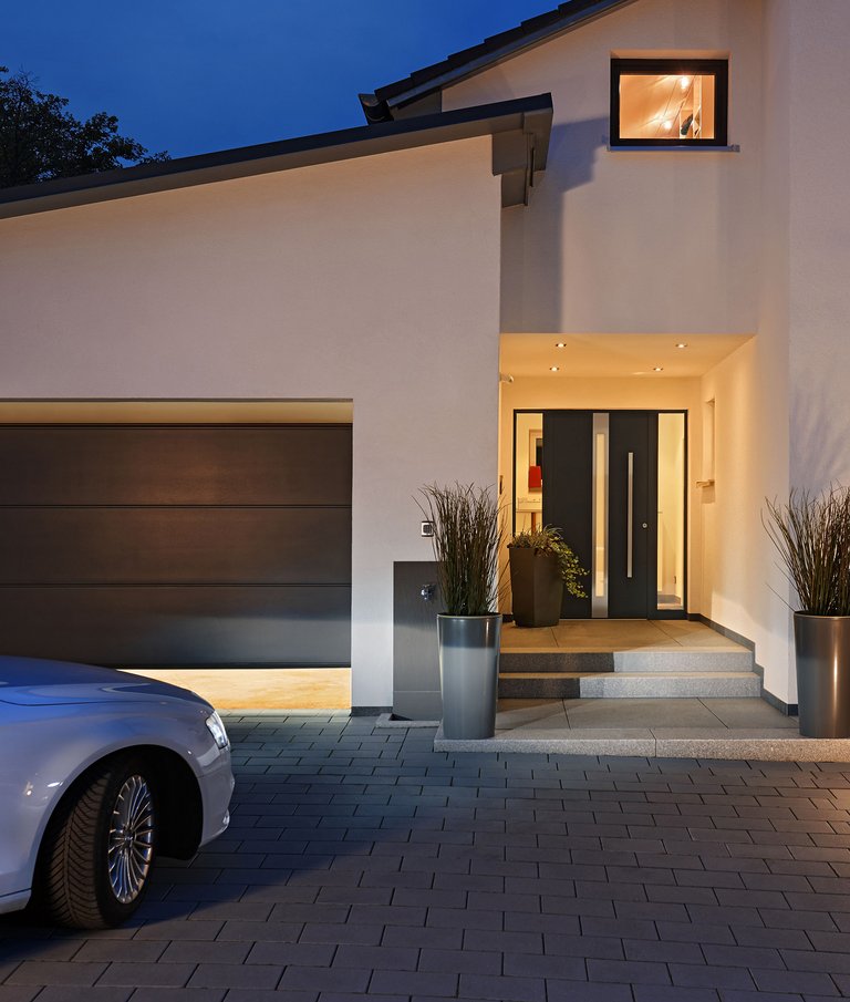 Einfahrt eines Hauses, in dem ein Auto in eine sich öffnende Garage fährt. Rechts Hauseingang in der Abenddämmerung