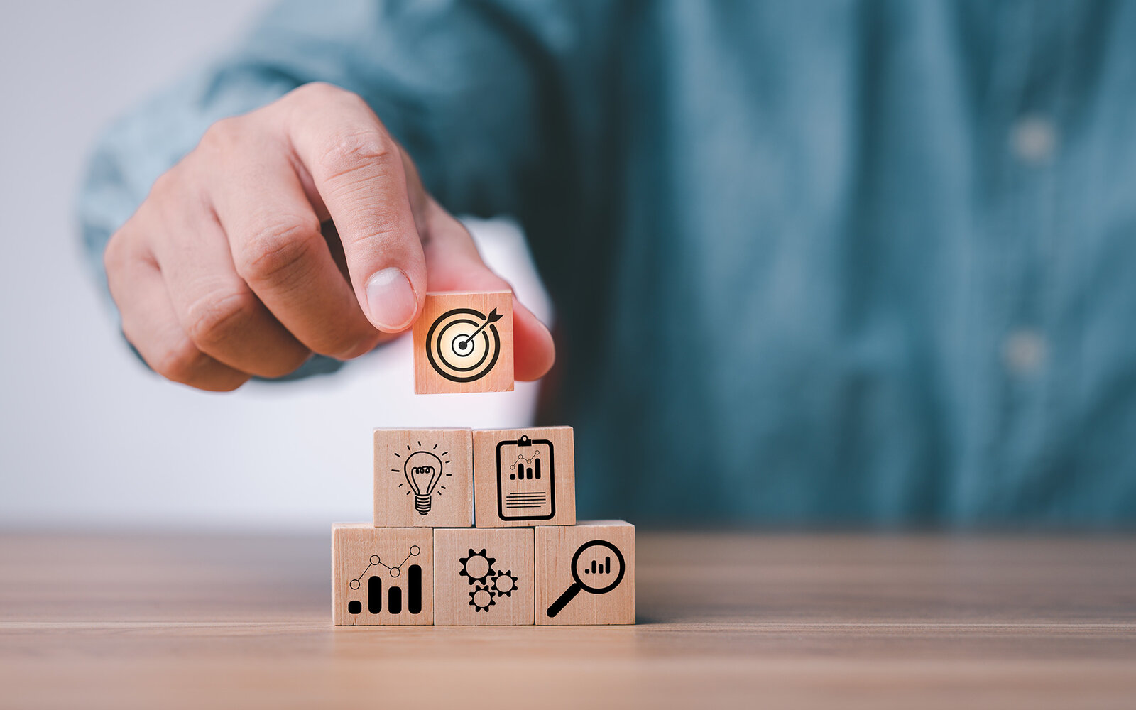 Ein Mann in blauem Hemd hält einen Holzbaustein mit einer Zielscheibe darauf in der Hand und legt sie auf 5 andere Bausteine, die aufeinander gestapelt sind