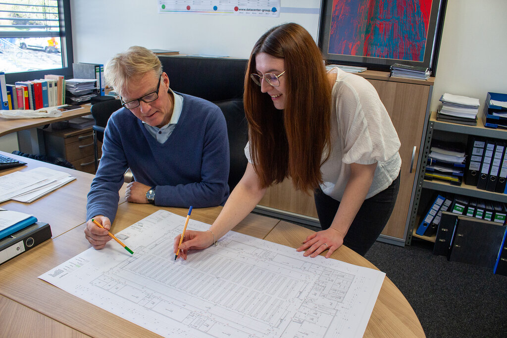 Eine Frau rechts steht und ein Mann links sitzt vor einem Schreibtisch, auf dem ein Plan liegt. Beide haben einen Stift in der Hand und zeigen auf den Plan