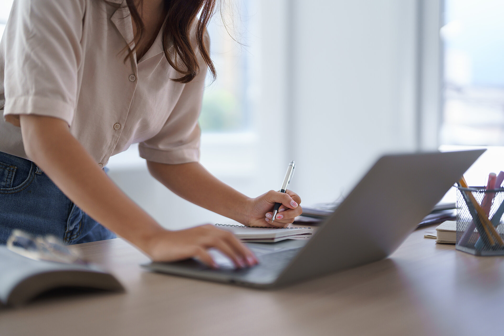 Dame steht an einem Schreibtisch, schaut auf einen Laptop und erstellt parallel Notizen auf einem Block