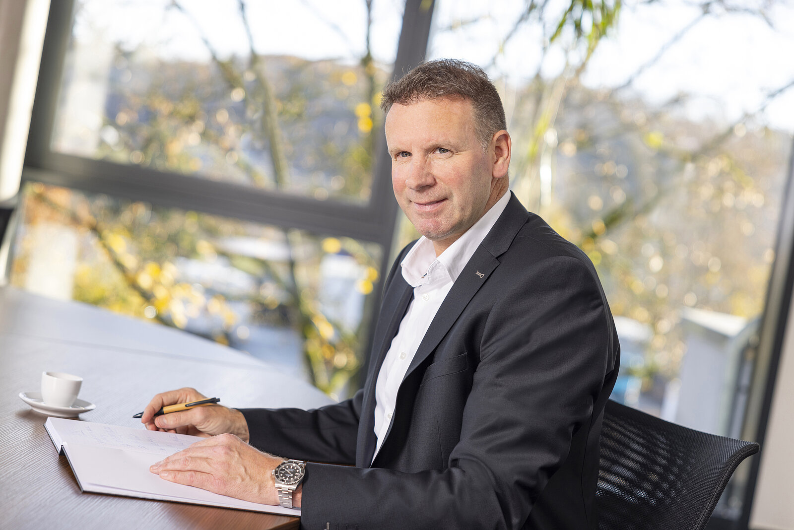 Ralf Siefen im Büro, im Anzug mit Armbanduhr und Stift in der Hand vor einem Buch und einer Kaffeetasse sitzend, setzt an zum schreiben