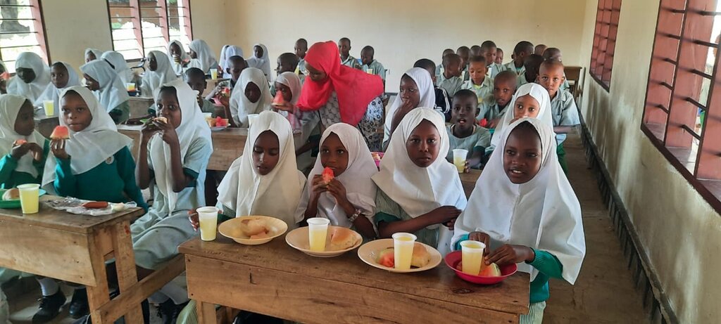 Kinder sitzen an Tischen im Klassenraum und essen Obst, Gebäck und trinken Limonade zur Eröffnungsfeier