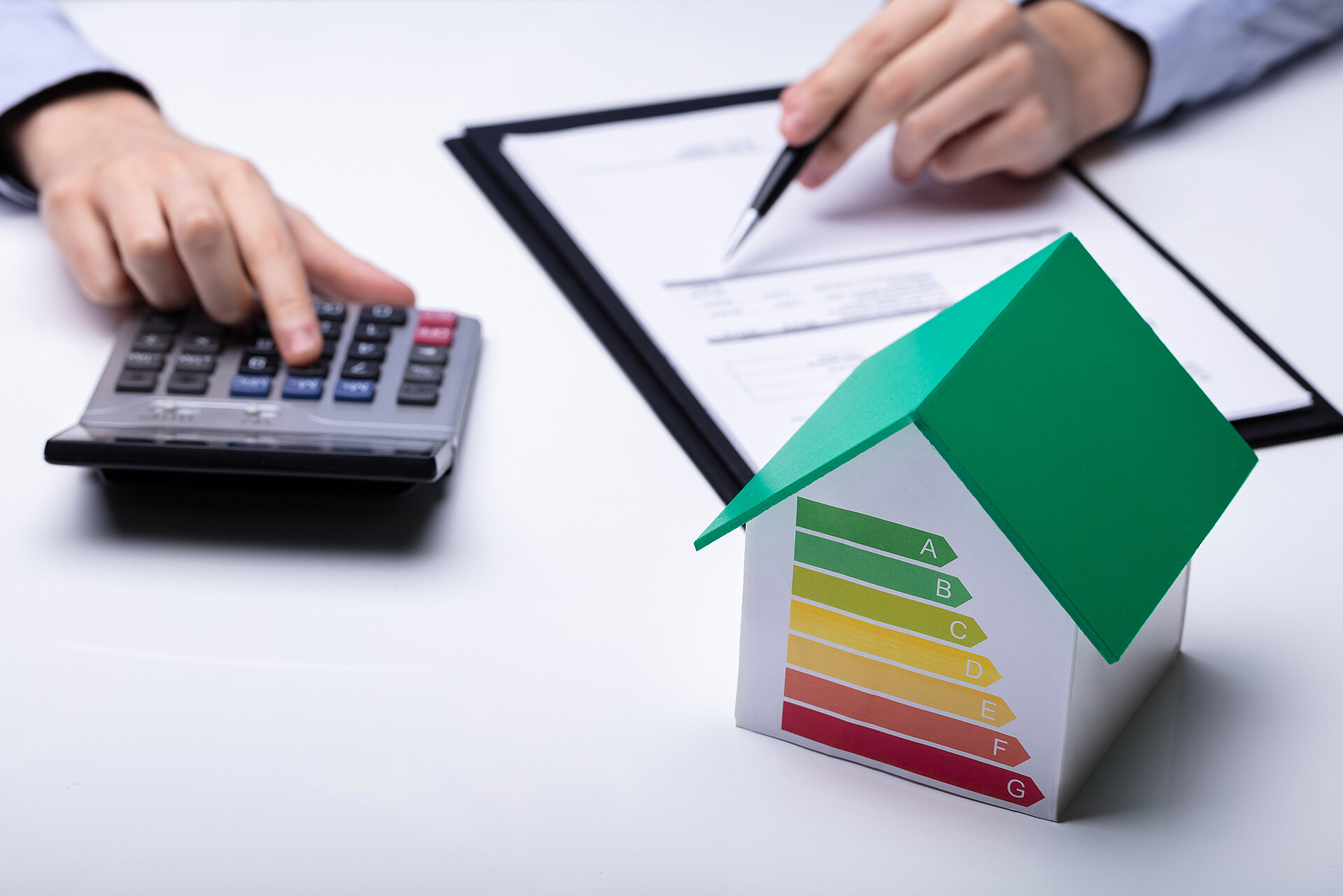 2 Hände, die einen Taschenrechner bedienen und schreiben. Dazu ein grünes Miniaturhaus