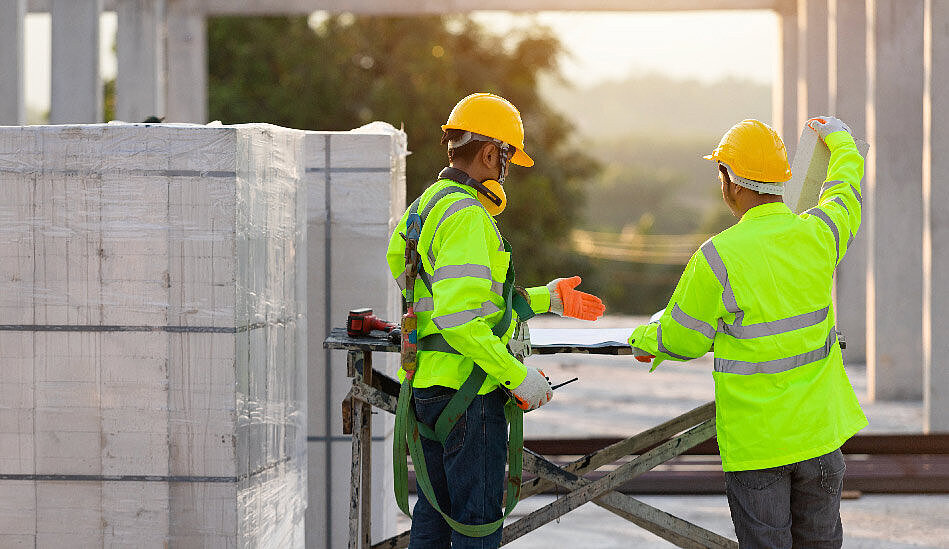 2 Männer verladen Baustoff. Beide Tragen Sicherheitskleidung, Helme, Handschuhe und Gehörschutz und Sicherheitsgurte