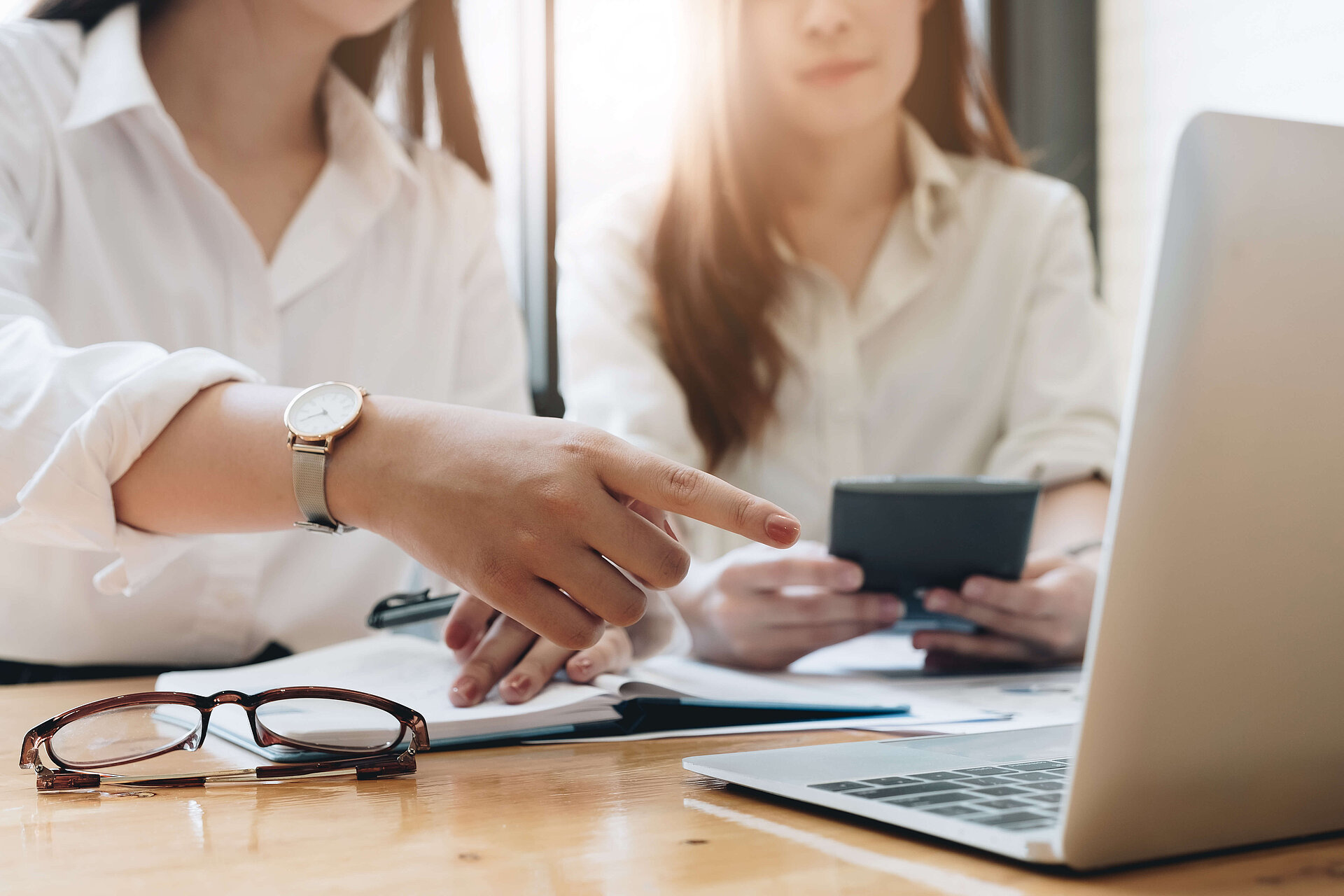 2 Frauen die an einem Schreibtisch sitzen. Eine Frau hält einen Taschenrechner in der Hand. Auf dem Schreibtisch befindet sich Papier, ein Laptop, auf den die andere Frau zeigt, und eine Brille