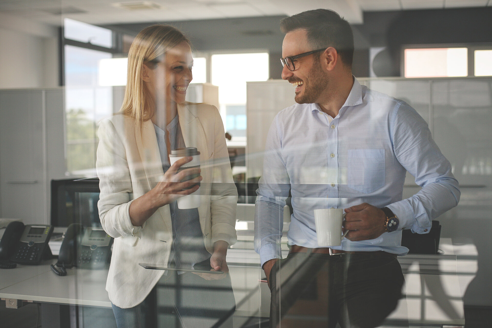 Mann und Frau in einem Büro vor hinter einer Glasfront, mit einer Tasse in der Hand, sich anlächelnd