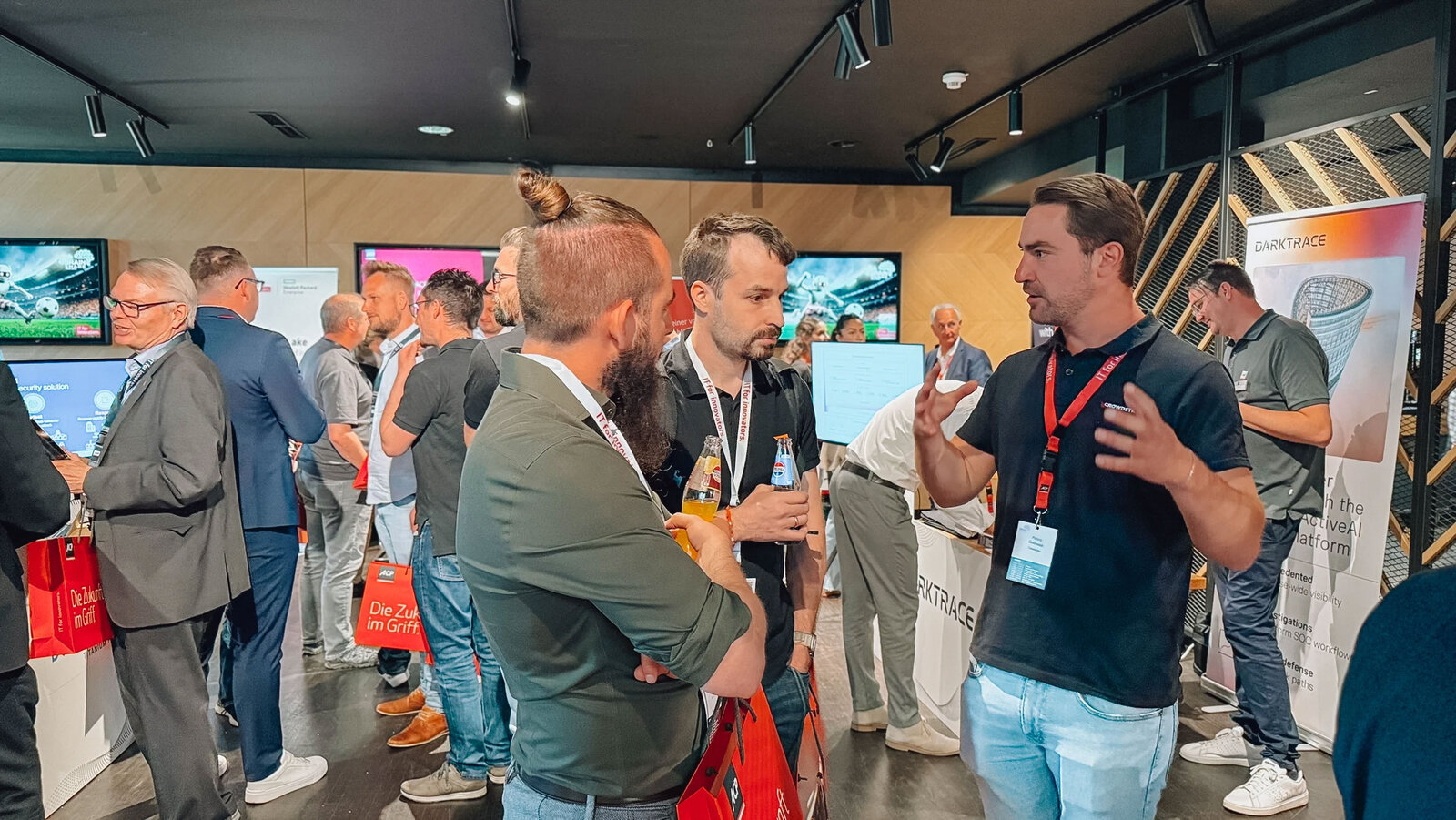 Menschen sich unterhaltend auf einer Messe, mit Getränken inder Hand, links ein BIldschirm