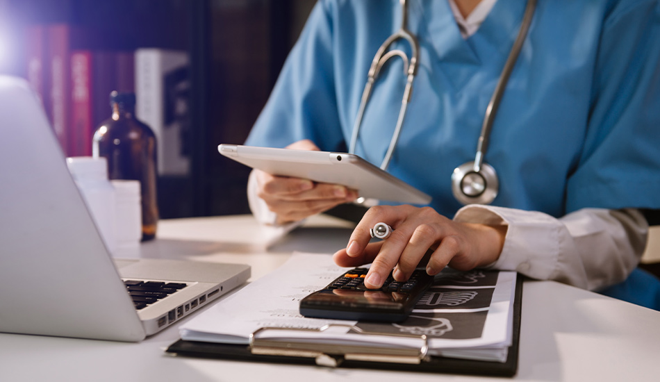 A doctor at a desk with a stethoscope around his neck. He is transferring something from a tablet to a hand. Underneath lies a clipboard. Next to it is a laptop