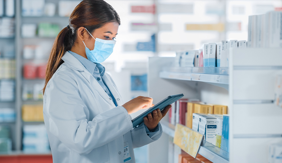 A woman wearing a mouth guard in a pharmacy works on a tablet. In the background a cabinet full of medicines