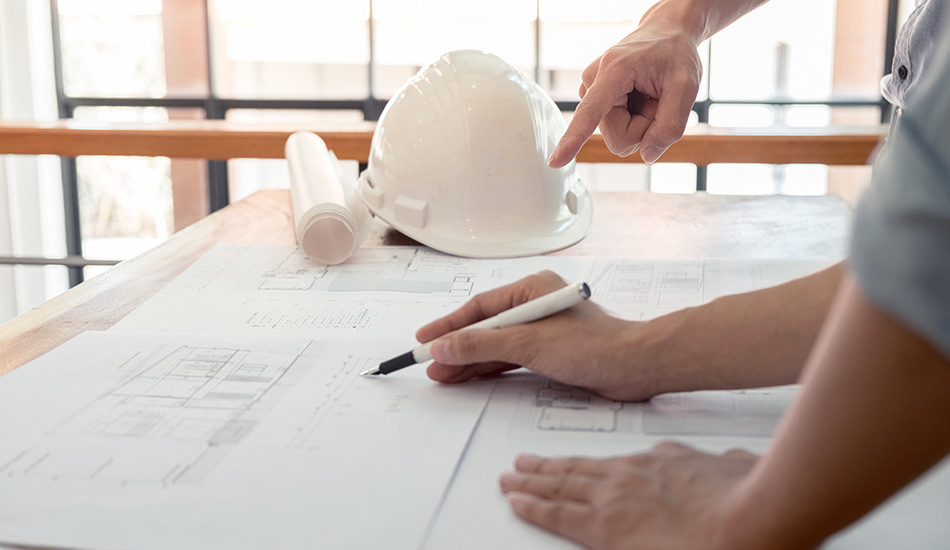 A plan on a desk with one man drawing something and the other pointing and hinting at the plan. On the desk is also a construction site helmet