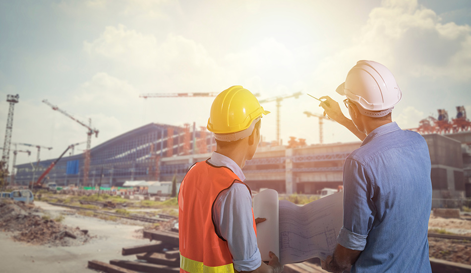 2 men with helmets are standing in front of a construction site with a crane. The right man points with his hand to the construction site