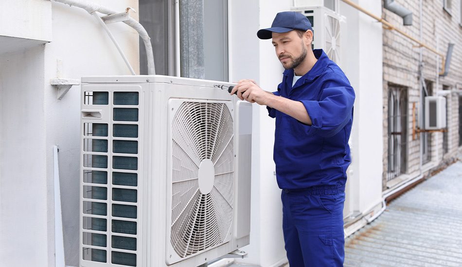 Mann mit blauer Arbeitskleidung und Kappe arbeitet mithilfe eines Schraubenziehers an einer Klimaanlage