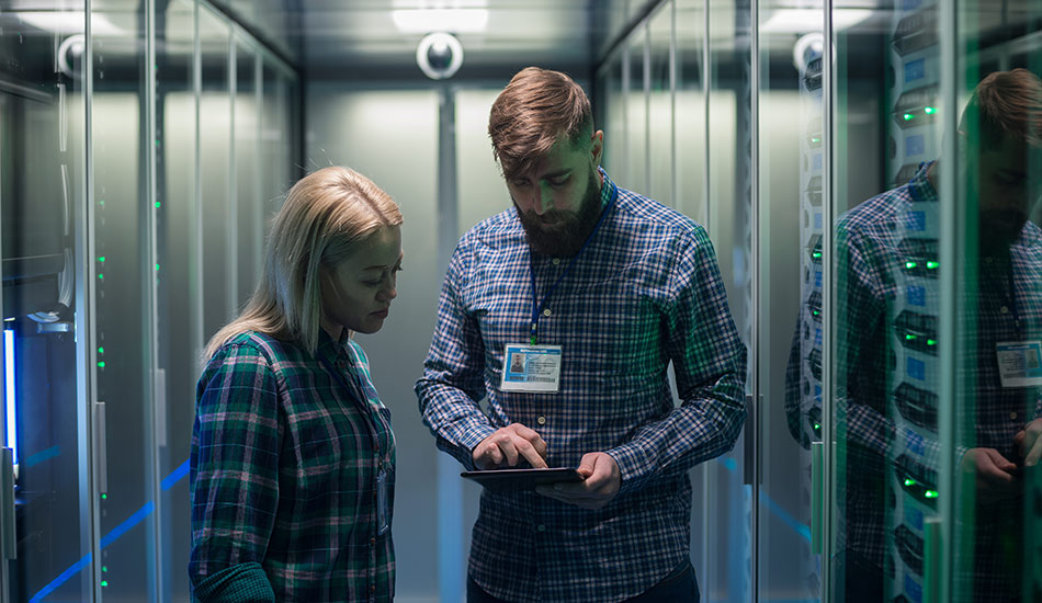 A man and a woman are standing in a data center. Both are looking at a mobile device