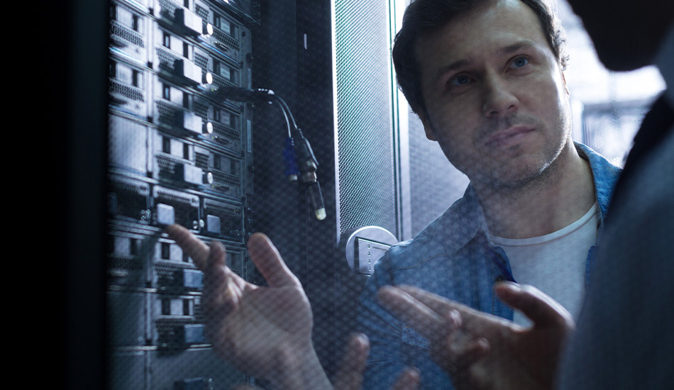 2 men are standing in front of a data center rack. Man 1 smiles and gestures to man 2 showing something