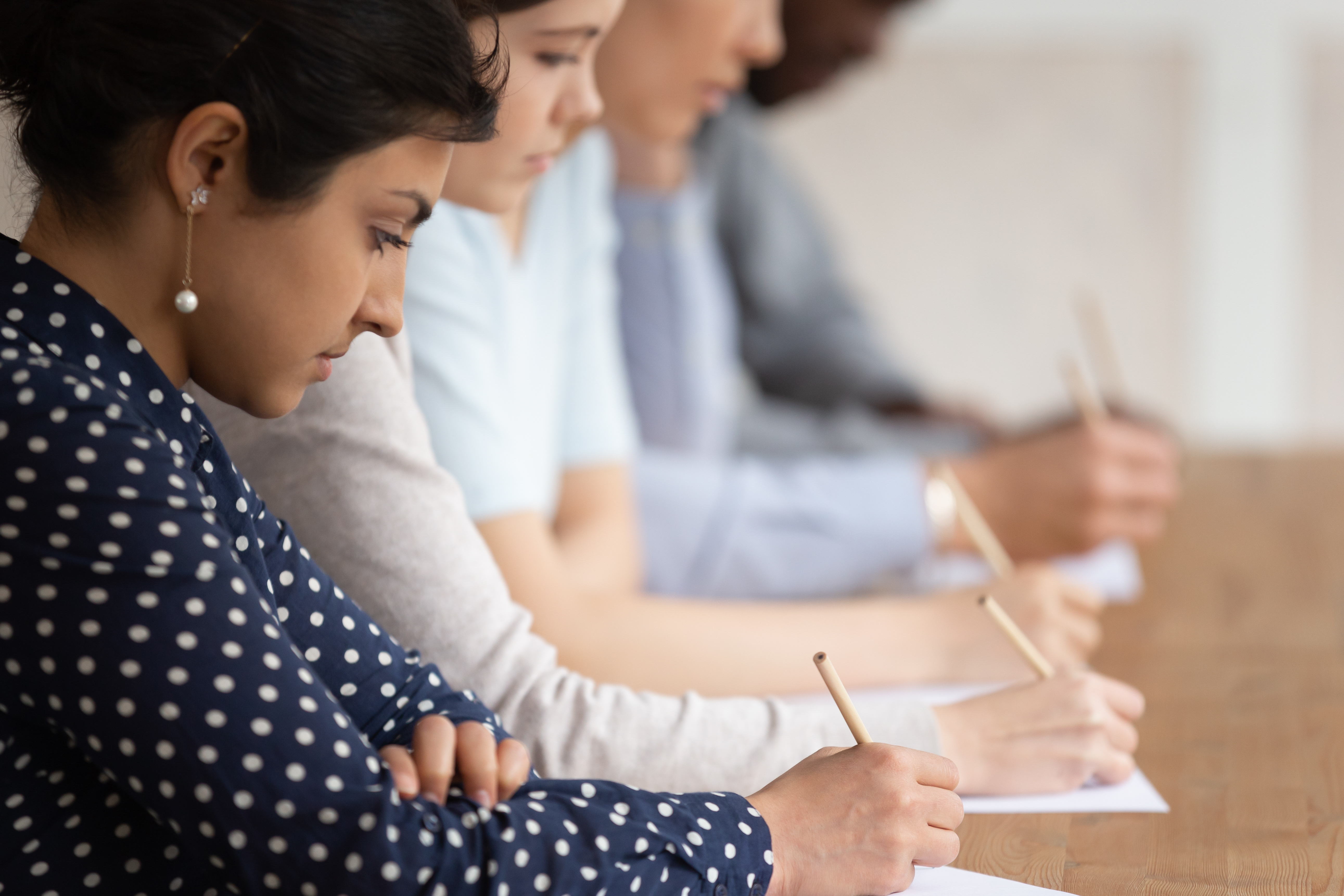 Studierende sitzen in einer Reihe an einem Schreibtisch und schreiben mit Bleistiften auf Papier