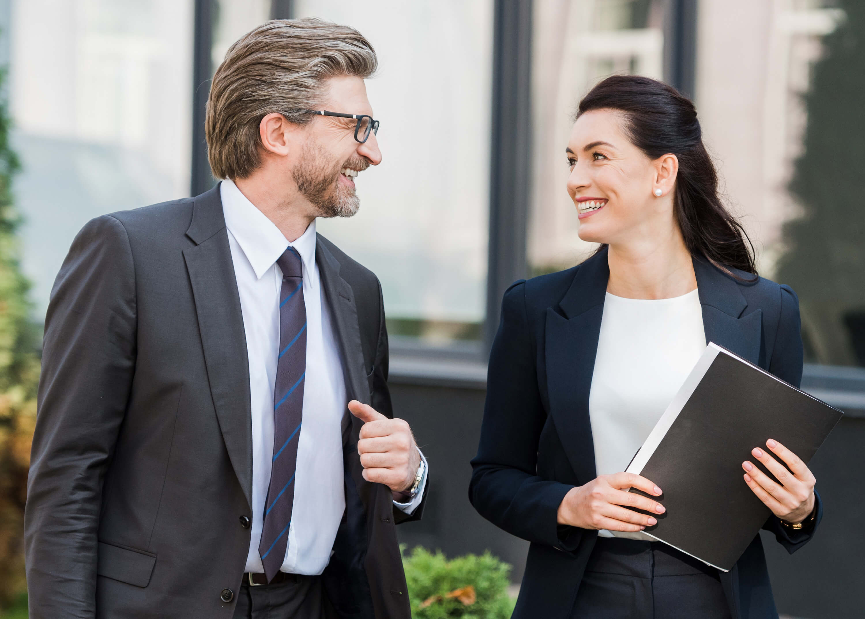 Man and woman smiling at each other, standing. Woman has a folder in her hand
