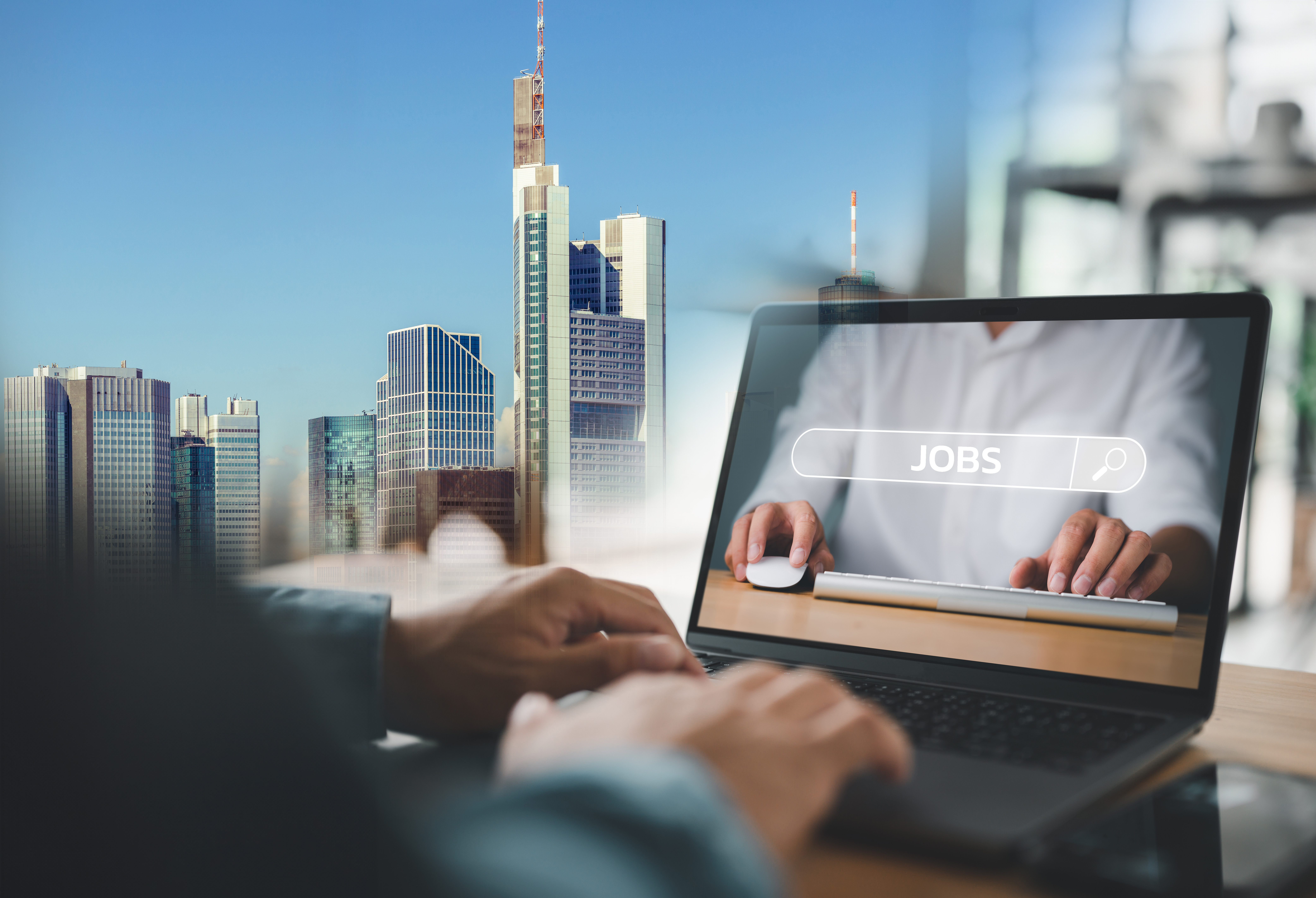 Im Vordergrund Mann, der an einem Laptop arbeitet und auf dem JObs steht, dahinter die Skyline von Frankfurt am Main