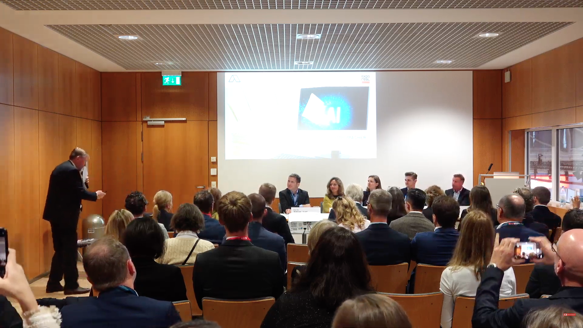Podiumsdiskussion auf der EXPO REAL mit Zuschauern und Panel, dahinter Präsentation auf einem Bildschirm