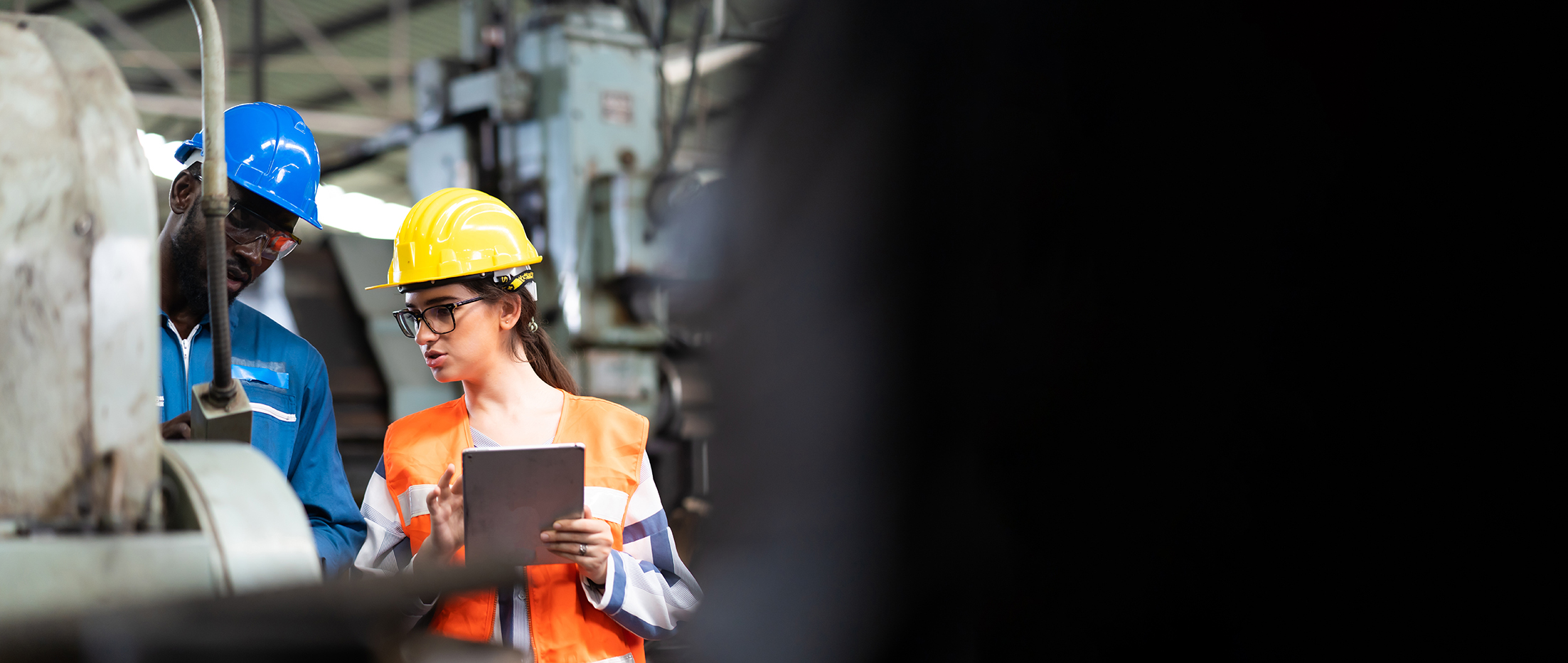 Mann mit blauer Montur und Frau mit gelbem Helm und orangener Weste blicken auf eine Maschine. Die Frau hält ein Klemmbrett in der Hand