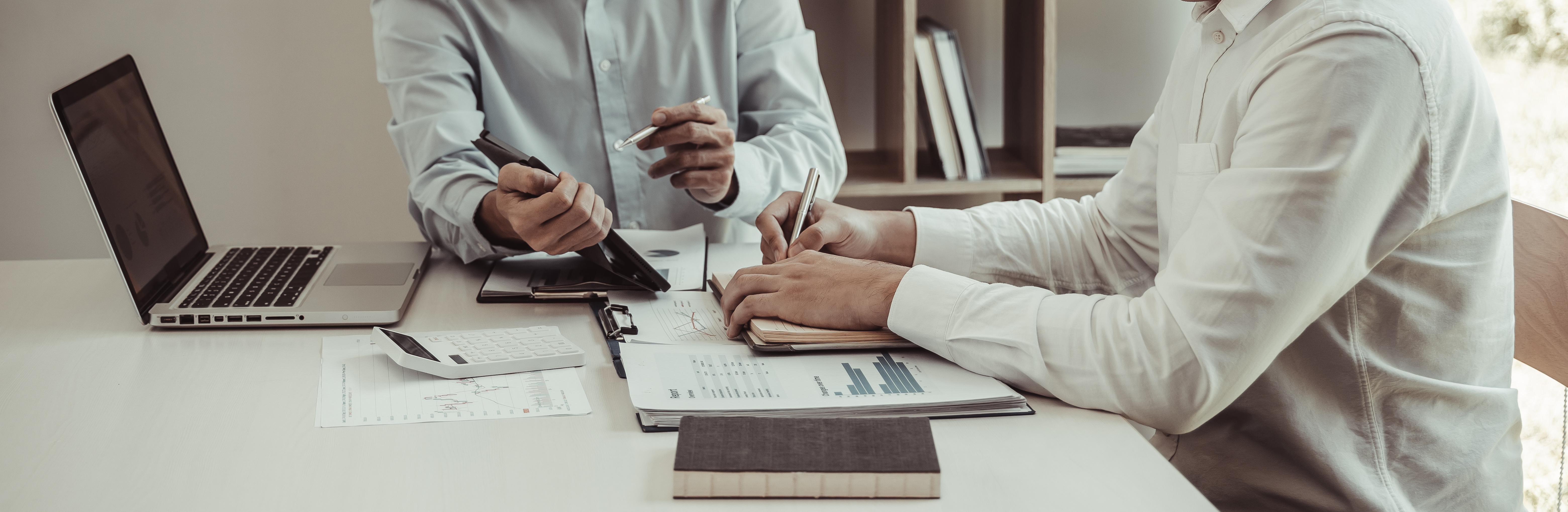 2 Männer an einem Schreibtisch. Mann 1 erklärt Mann mithilfe eines Stiftes an einem Laptop etwas