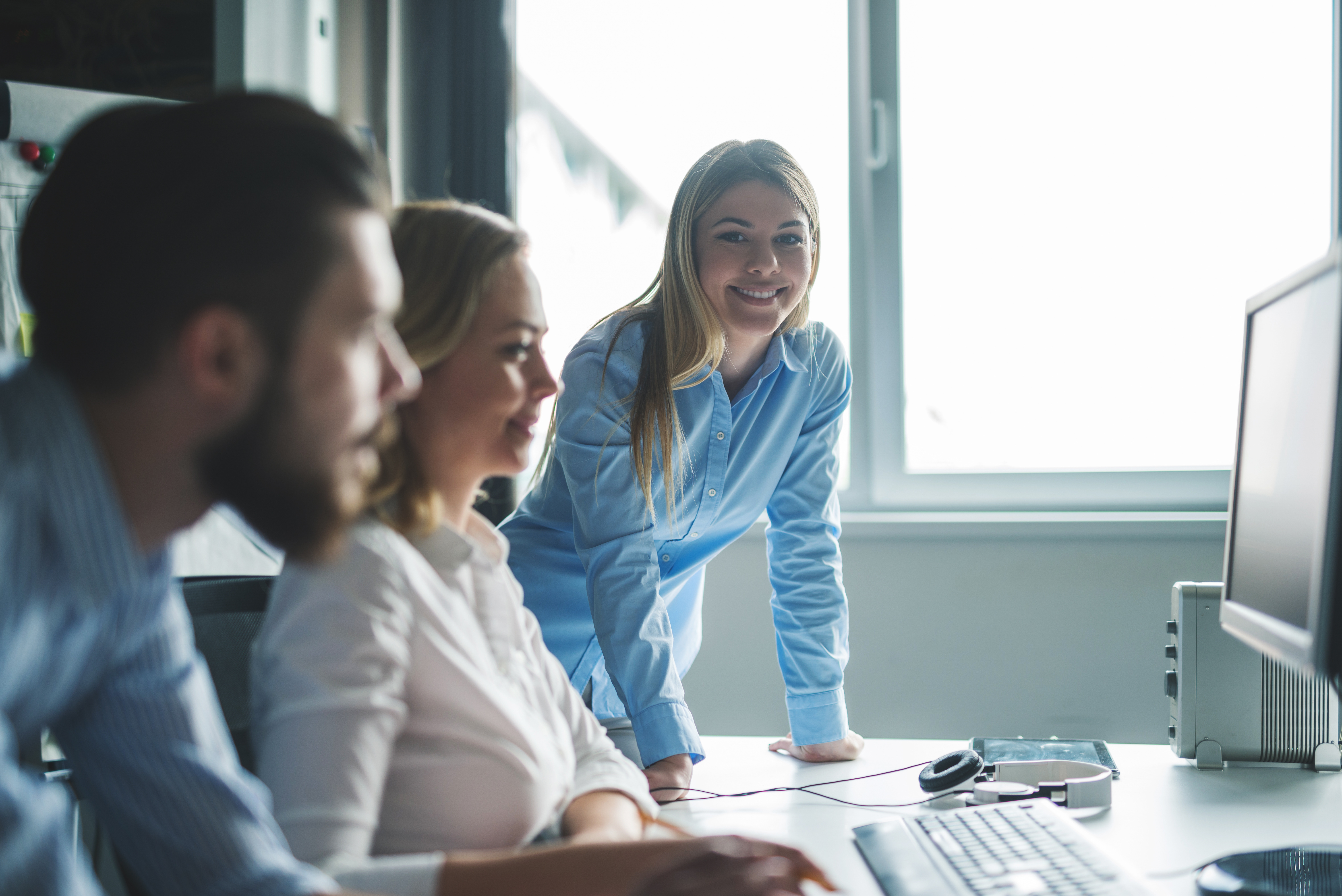 2 Frauen und ein Mann, die an einem Schreibtisch sitzen. Mann und eine Frau blicken auf einen Bildschirm, eine Frau schaut in die Kamera